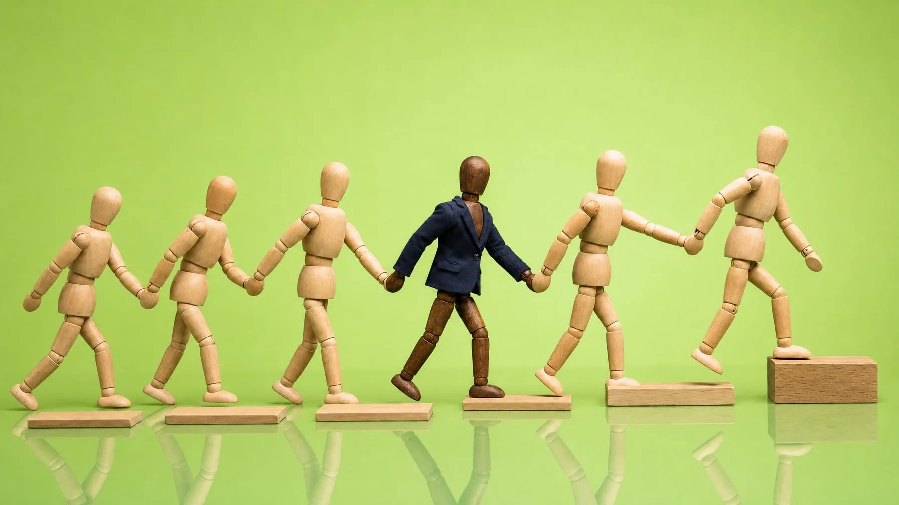 Six wooden mannequins walk hand in hand from left to right over flat wooden steps, the first one climbing the highest, a mannequin in a blazer in the middle of the chain